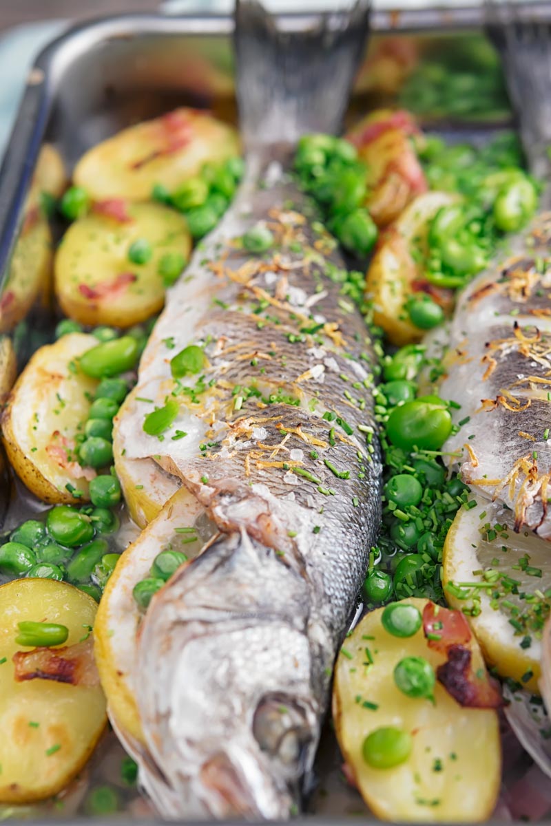 Portrait image of whole baked sea bass in a tray with potatoes peas and broad beans