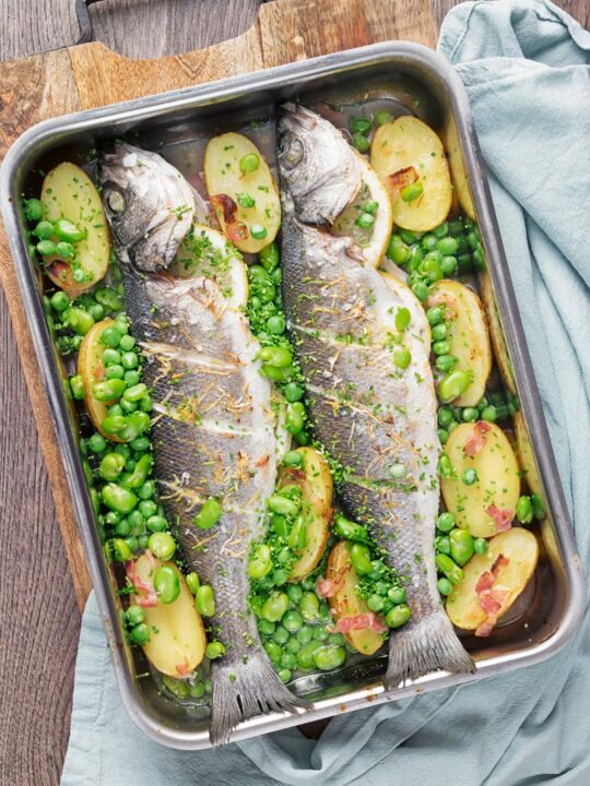 Portrait overhead image of two whole baked sea bass in a tray with potatoes peas and broad beans