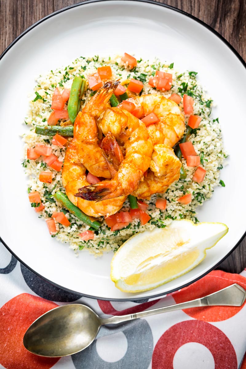 Portrait overhead image of harissa garlic prawns served on a bed of buttered couscous served with a lemon wedge on a white plate