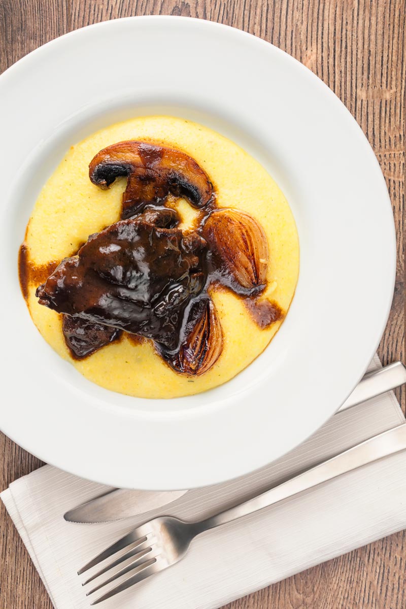Portrait overhead image of beer braise beef cheeks with a rich glaze served with seared shallots and mushrooms on polenta