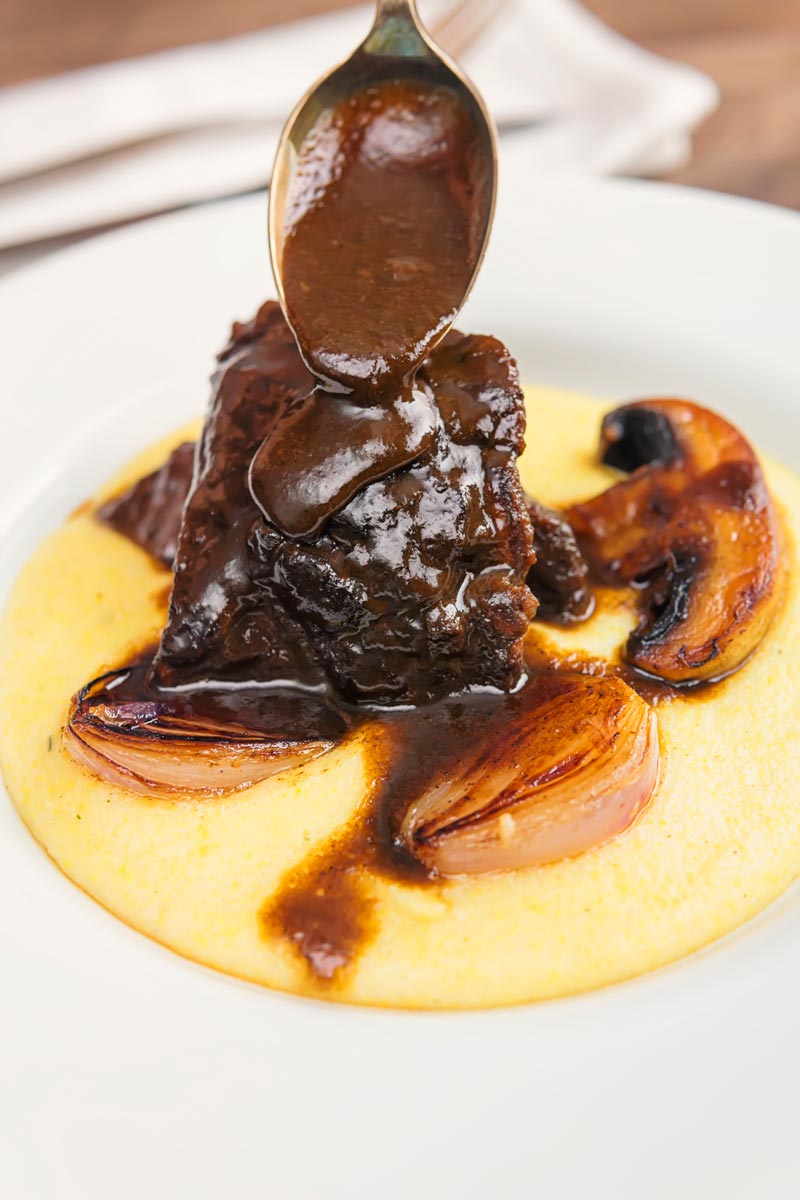 Portrait image of beer braised beef cheeks being glazed with a rich sauce served with seared shallots and mushrooms on polenta