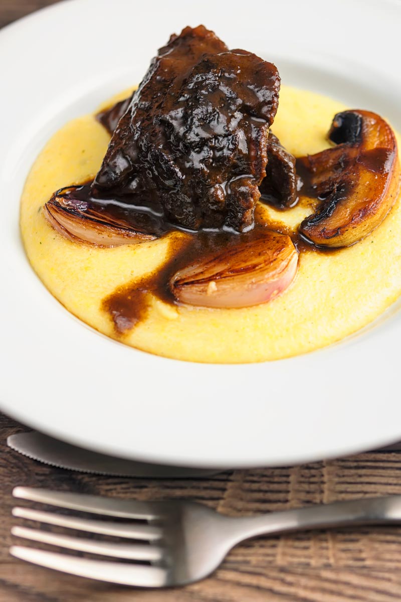 Portrait image of beer braised beef cheeks with a rich glaze served with seared shallots and mushrooms on polenta