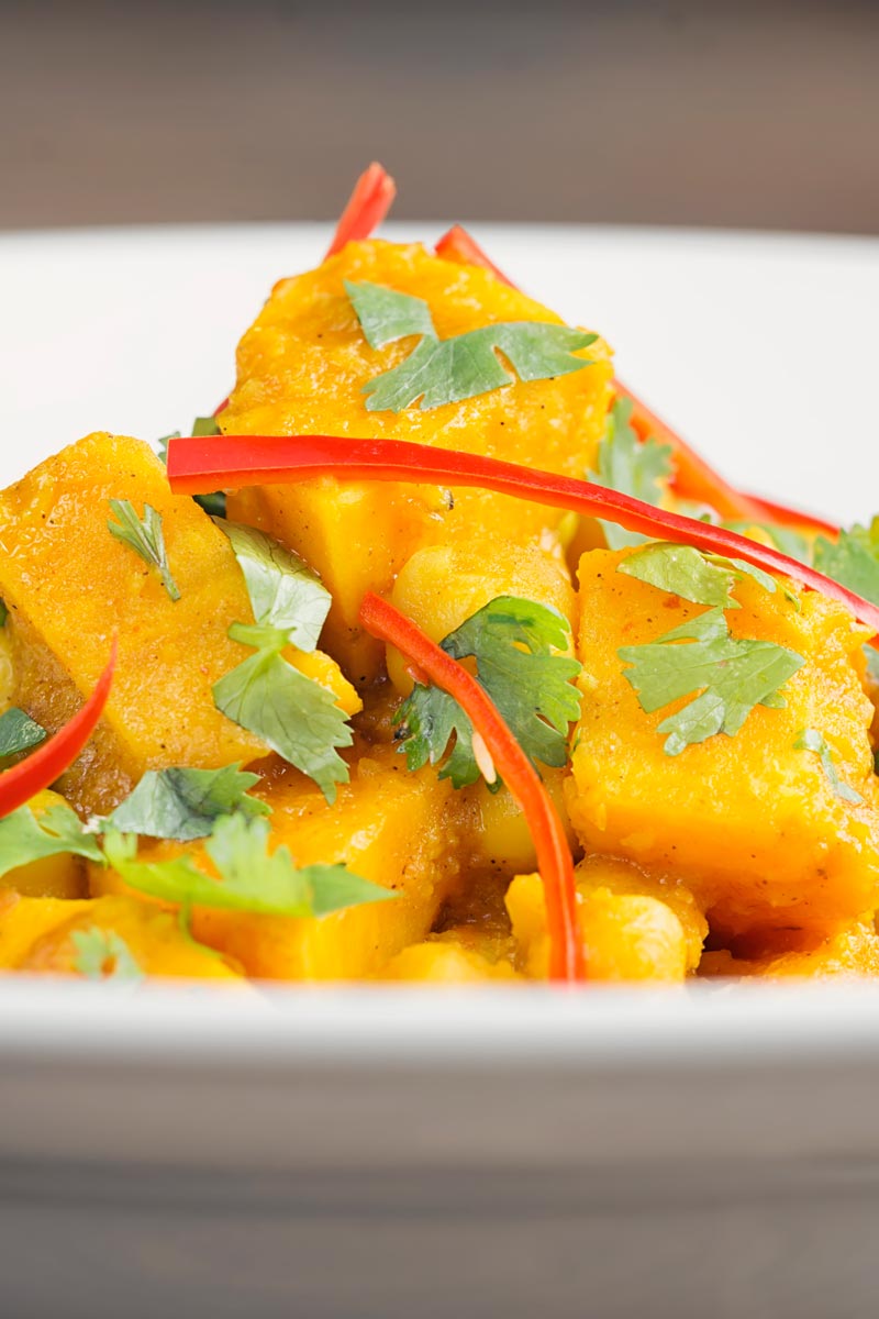 Portrait close up picture of a golden chickpea and sweet potato curry with a white bowl with chopped coriander and shredded chilli