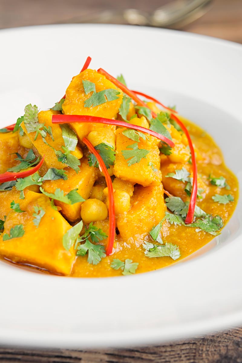 Portrait picture of a golden chickpea and sweet potato curry with a white bowl with chopped coriander and shredded chilli