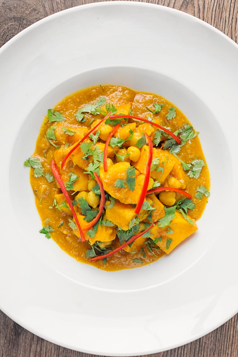 Portrait overhead picture of a golden chickpea and sweet potato curry with a white bowl with chopped coriander and shredded chilli