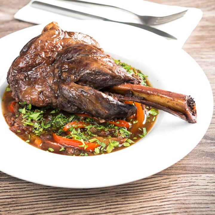 Square image of a Slow Cooker Lamb Shank served on a bed of braising liquid and red peppers served in a shallow white bowl