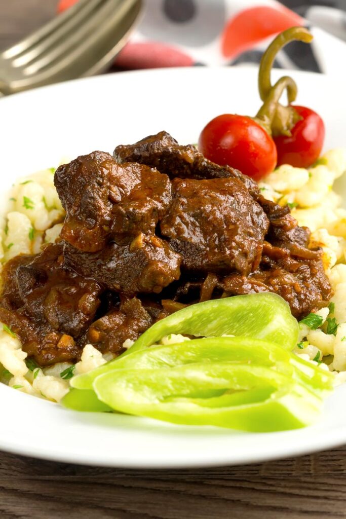 Portrait image of a marha porkolt, a Hungarian beef stew served with nokedli, hot wax peppers and pickled cherry bomb chilli peppers served in a white bowl