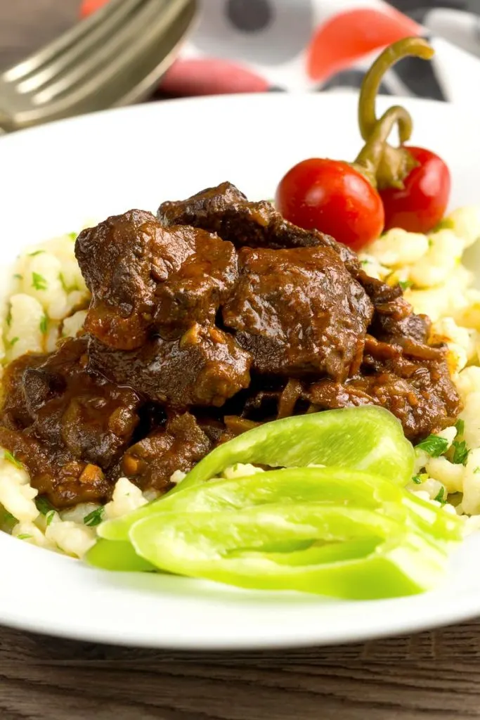 Portrait image of a marha porkolt, a Hungarian beef stew served with nokedli, hot wax peppers and pickled cherry bomb chilli peppers served in a white bowl