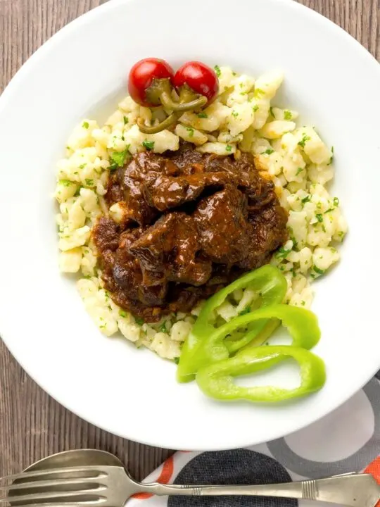 Portrait overhead image of a marha porkolt, a Hungarian beef stew served with nokedli, hot wax peppers and pickled peppers served in a white bowl