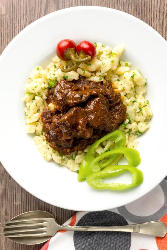 Portrait overhead image of a marha porkolt, a Hungarian beef stew served with nokedli, hot wax peppers and pickled peppers served in a white bowl
