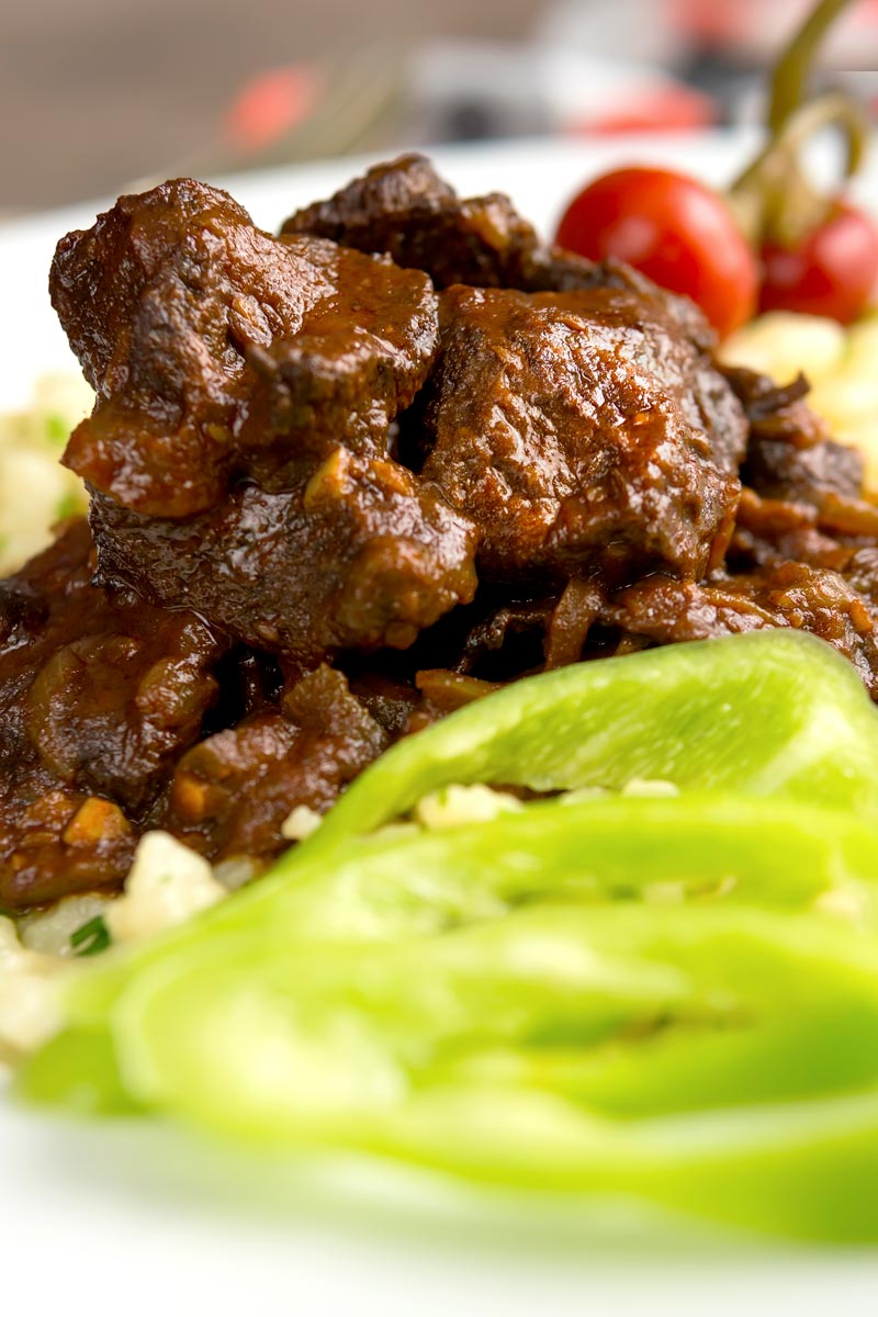 Portrait close up overhead image of a marha porkolt, a Hungarian beef stew served with nokedli, hot wax peppers and pickled peppers served in a white bowl