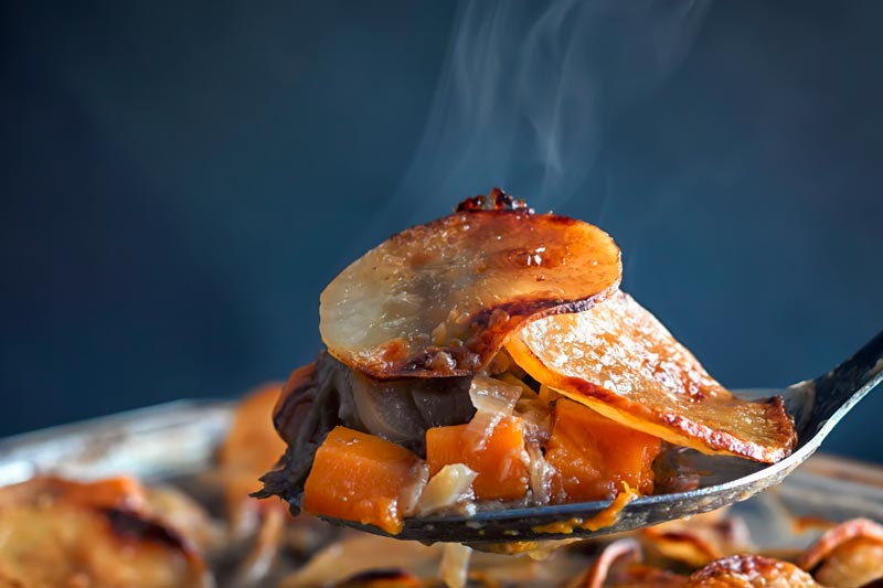 Landscape image of a steaming portion of lamb hot pot being taken from a pie featuring sliced potatoes and carrots