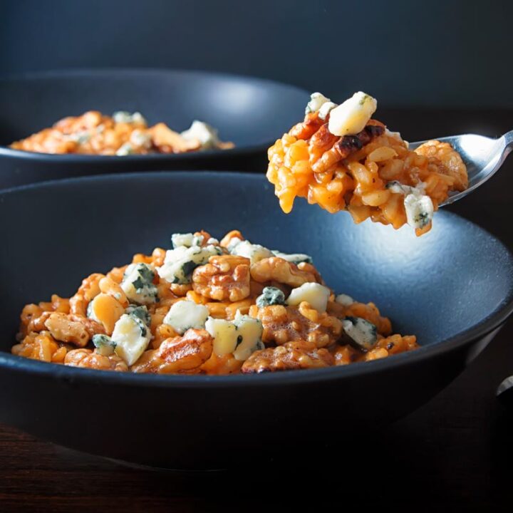 Square image of a pureed pumpkin risotto with blue cheese and walnuts served in a black bowl with spoon taking a portion
