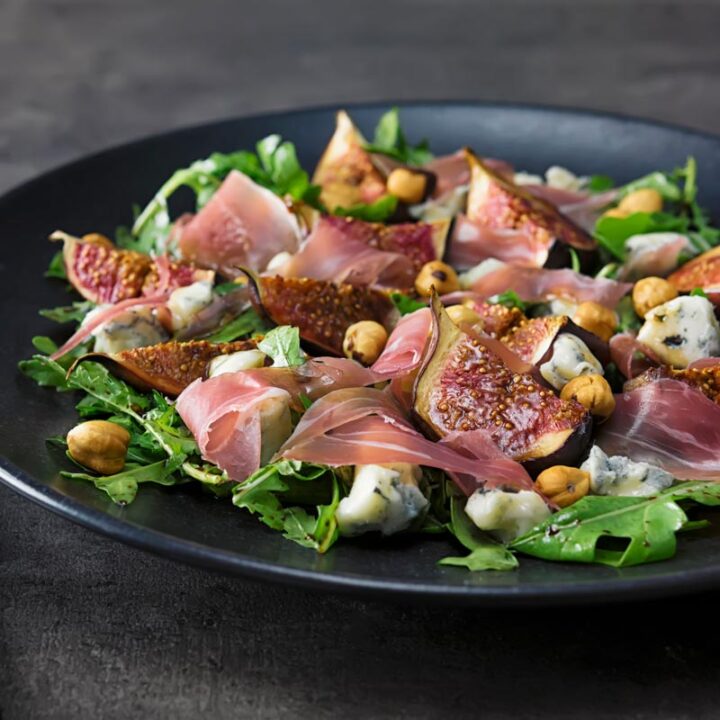 Square image of a roasted fig salad paired with blue cheese , rocket, Prosciutto Ham and hazelnuts served on a black plate