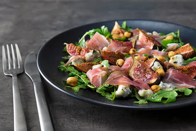 Landscape image of a roasted fig salad paired with blue cheese, rocket, Prosciutto Ham and hazelnuts served on a black plate