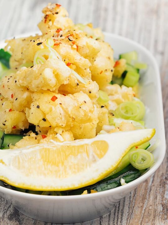 Portrait image of crispy salt and pepper squid served on pickled cucumber , sliced spring onions and a wedge of lemon served in a shallow white bowl