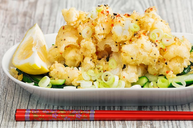 Landscape image of crispy salt and pepper squid served on pickled cucumber , sliced spring onions and a wedge of lemon served in a shallow white bowl