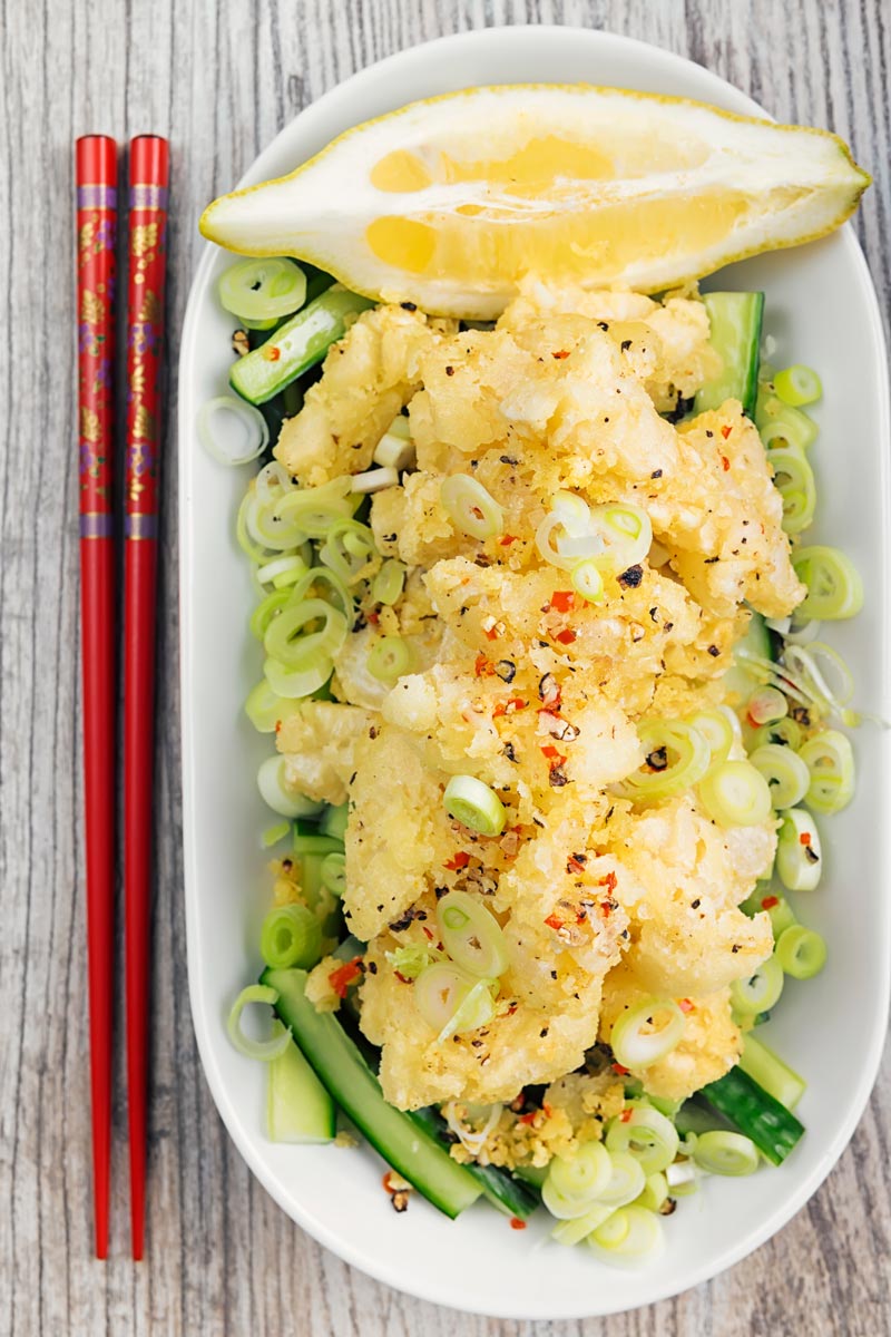 Portrait overhead image of crispy salt and pepper squid served on pickled cucumber, sliced spring onions and a wedge of lemon served in a shallow white bowl