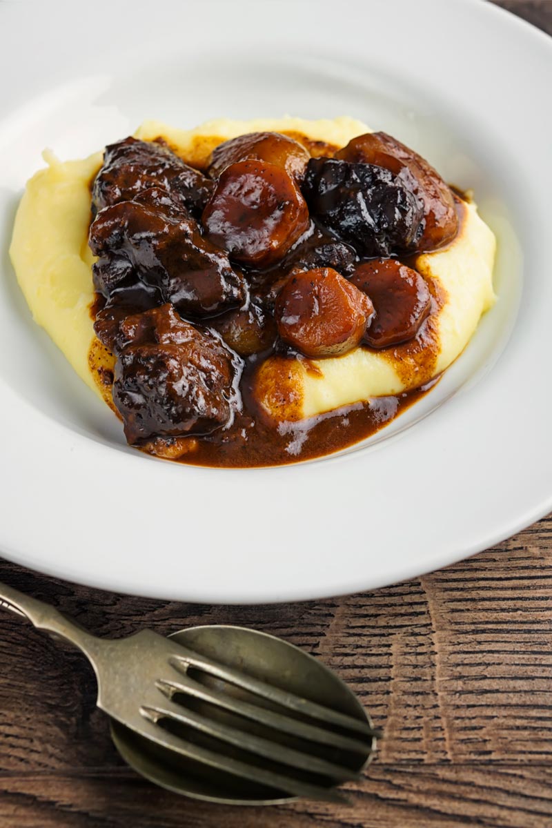 Portrait image of a daube of beef stew on a bed of creamy mashed potato served in a shallow white bowl