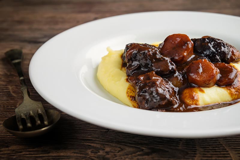 Landscape image of a daube of beef stew on a bed of creamy mashed potato served in a shallow white bowl