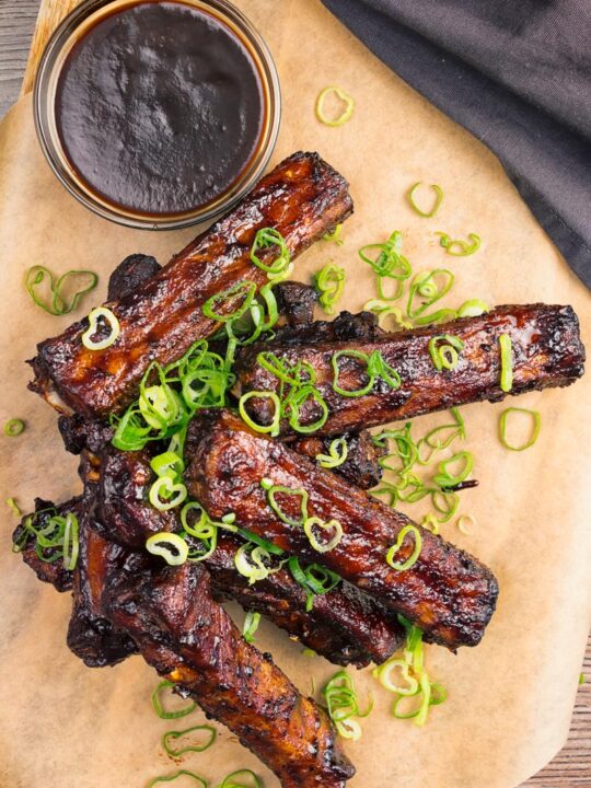 Portrait overhead image of a pile of sticky Instant Pot Chinese Spare Ribs served on a board with spring onions and a dipping sauce