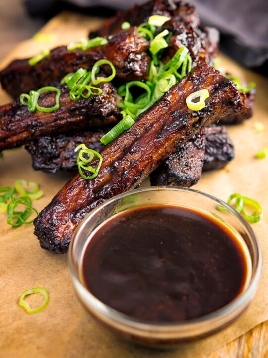 Portrait image of a pile of sticky Instant Pot Chinese Spare Ribs served on a board with spring onions and a dipping sauce