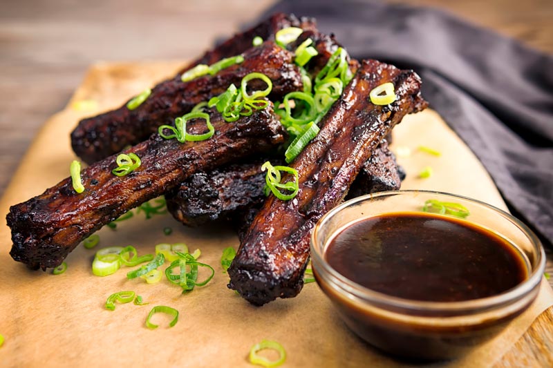 Landscape image of a pile of sticky Chinese Spare Ribs served on a board with spring onions and a dipping sauce