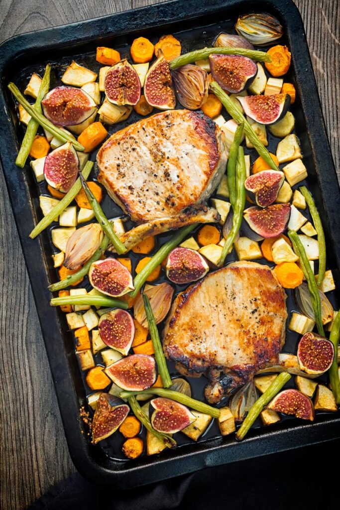 Portrait overhead image of a pork chop tray bake featuring fresh figs, green beans, shallots, carrots and parsnips