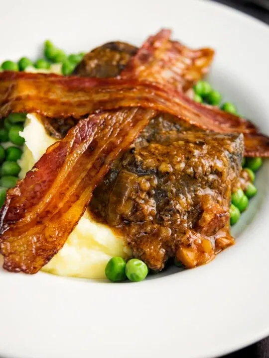 Portrait image of bacon liver an onions with mash and peas served in a white bowl