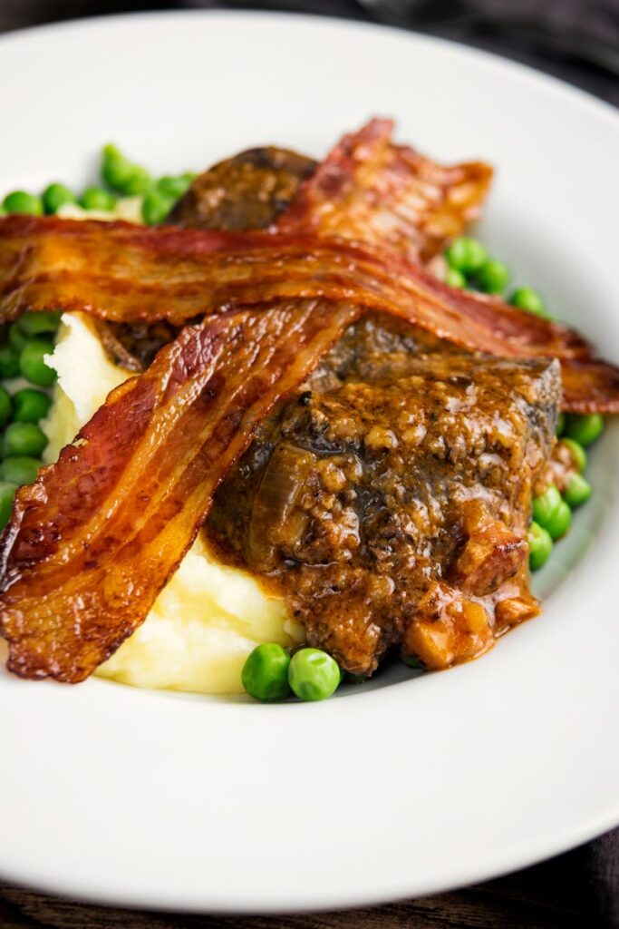 Portrait image of bacon liver an onions with mash and peas served in a white bowl