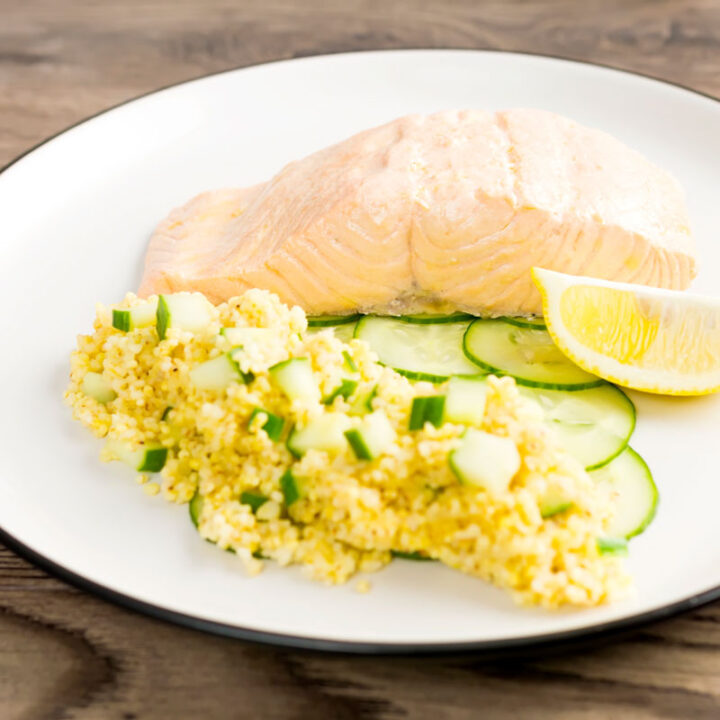 Square image of a poached salmon fillet served with cucumber and millet on a white plate with a wedge of lemon