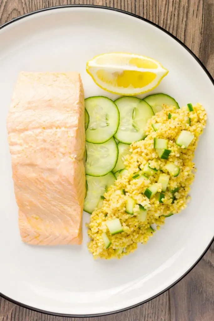Portrait overhead image of a poached salmon fillet served with cucumber and millet on a white plate with a wedge of lemon