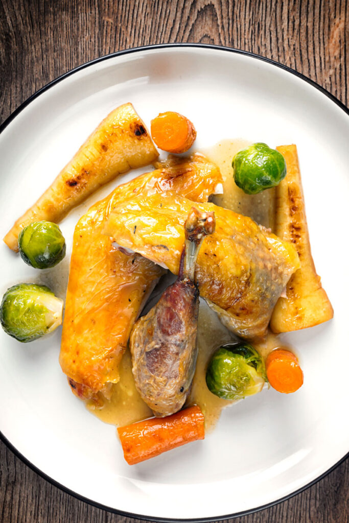 Portrait overhead image of a guinea fowl leg served on a white plate with honey glazed vegetables