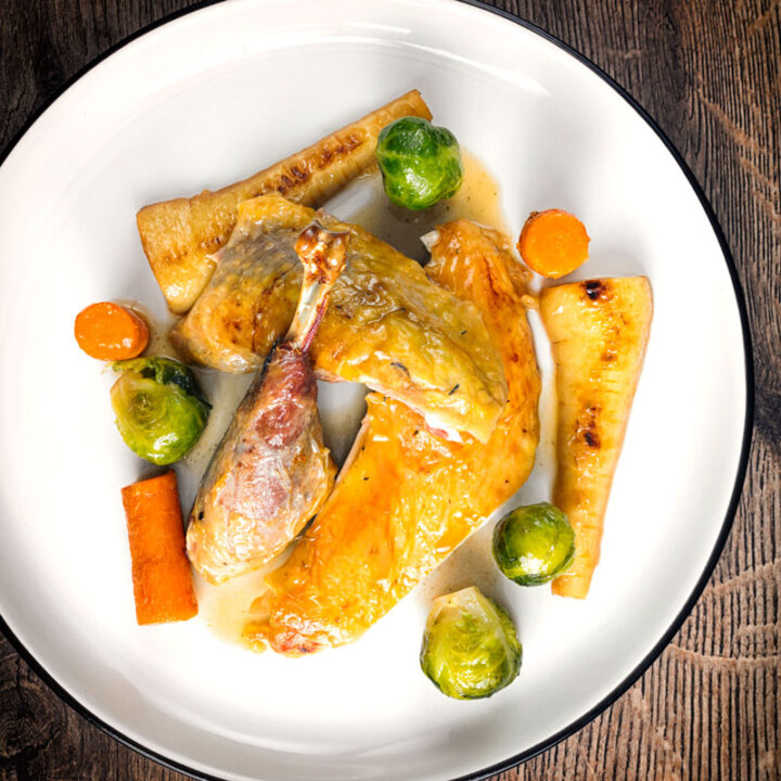 Square overhead image of a roast guinea fowl leg served on a white plate with honey glazed vegetables