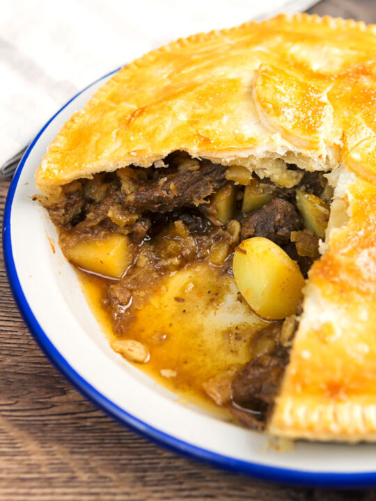 Portrait image of a meat and potato pie cooked in an enamel plate with a slice removed showing pie filling