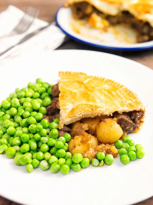 Portrait image of a slice of meat and potato pie served with garden peas on a white plate with the pie out of focus behind