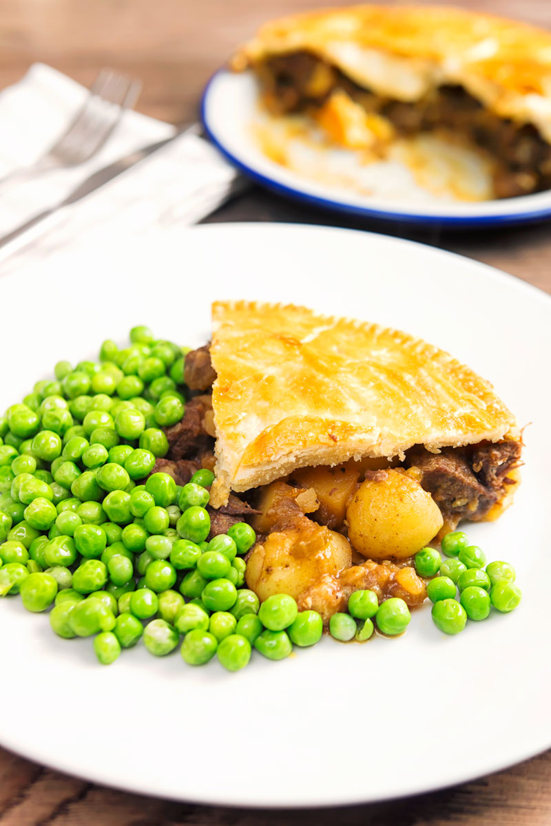 Portrait image of a slice of meat and potato pie served with garden peas on a white plate with the pie out of focus behind