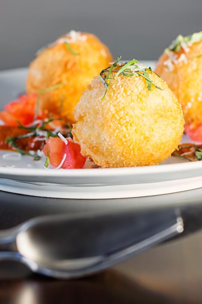Portrait close up image of three golden stuffed arancini balls served on a white plate with tomato sauce, tomato concase and shredded basil