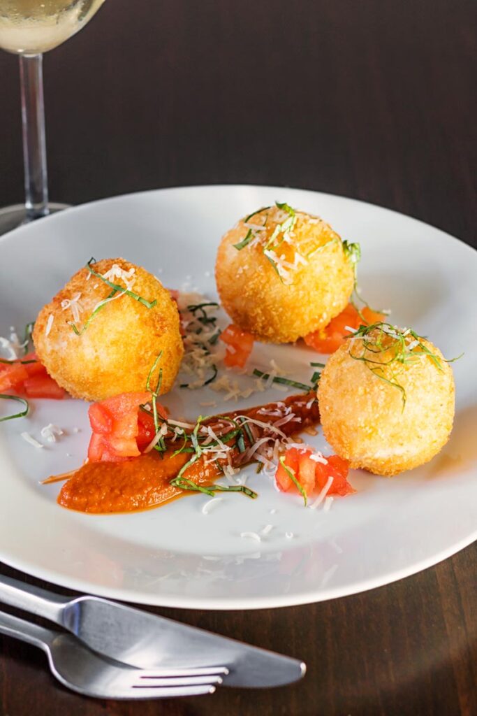 Portrait image of three golden stuffed arancini balls served on a white plate with tomato sauce, tomato concasse and shredded basil