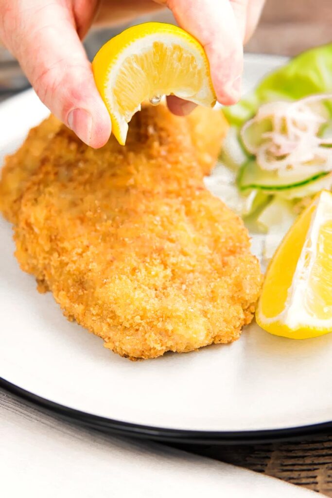 Portrait image of a lemon wedge being squeezed over a classic breaded veal Wiener schnitzel served with a classic green salad on a white plate
