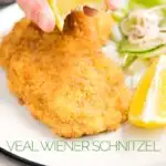 Portrait image of a lemon wedge being squeezed over a classic breaded veal Wiener schnitzel served with a classic green salad on a white plate with a text overlay