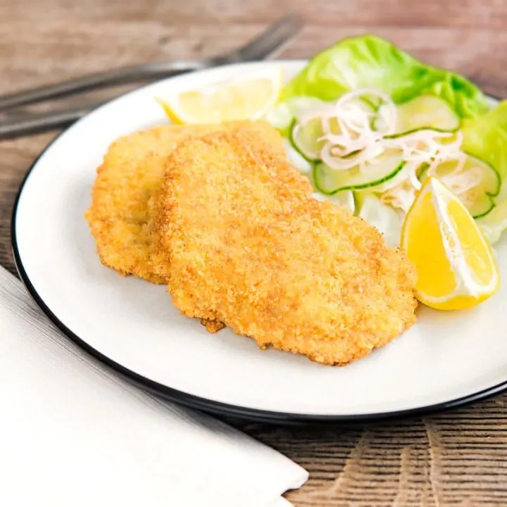 Square image of a classic breaded veal Wiener schnitzel served with a classic green salad on a white plate