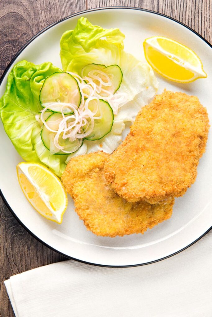 Portrait overhead image of a classic breaded veal Wiener schnitzel served with a classic green salad and lemon wedges on a white plate