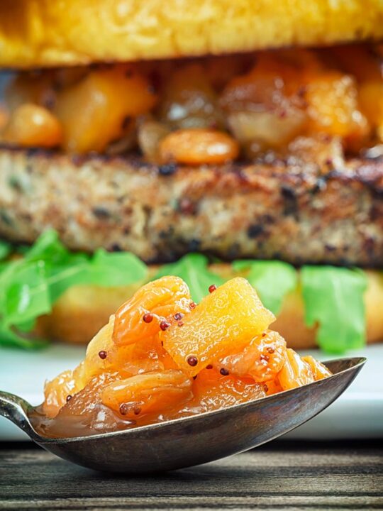 Close up portrait image of a spoonful of chunky apple chutney with an out of focus burger in the background
