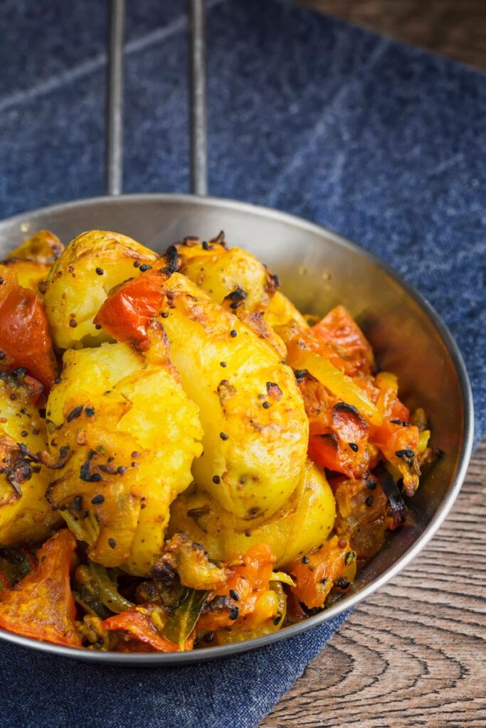 Portrait image of lightly crushed Bombay potatoes served in a small pan on a blue cloth