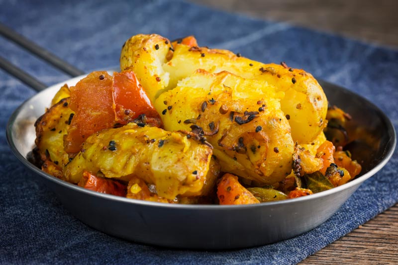 Landscape image of lightly crushed Bombay potatoes served in a small pan on a blue cloth
