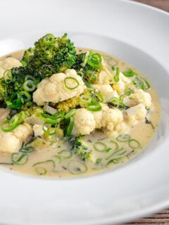 Portrait image of a green Thai curry soup with cauliflower and broccoli served in a white bowl