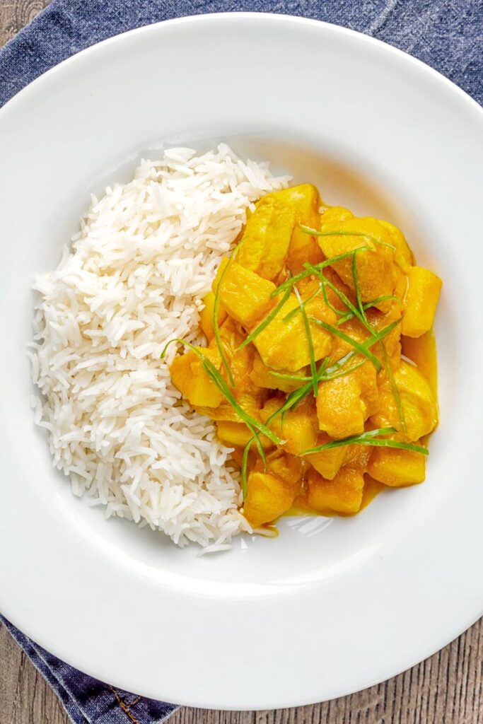 Portrait overhead image of a chicken and pineapple curry in a thick sauce with a chiffonade of green chilli and white rice served in a white bowl