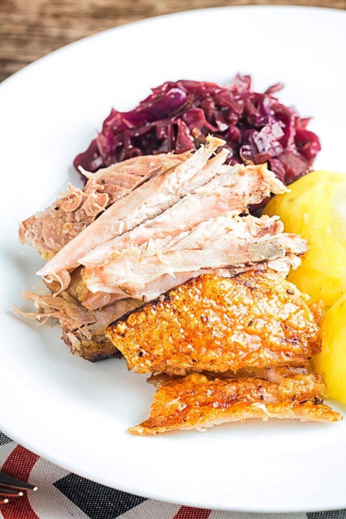 Portrait image of the meat from a roast pork knuckle served on a white plate with crispy crackling, red cabbage and boiled potatoes.