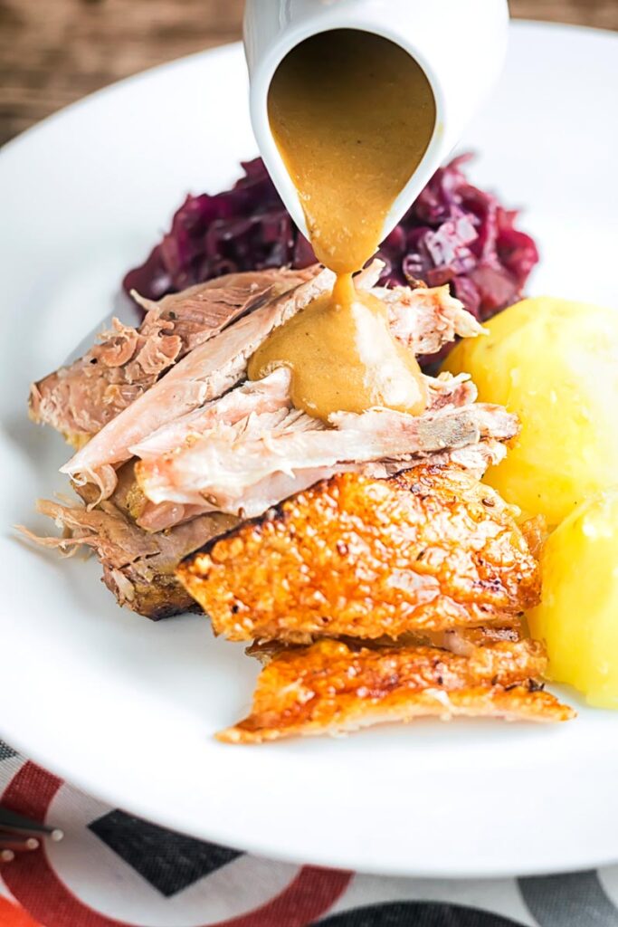 Portrait image of the meat from a roast pork knuckle having gravy poured over it served on a white plate with crispy crackling, red cabbage and boiled potatoes.